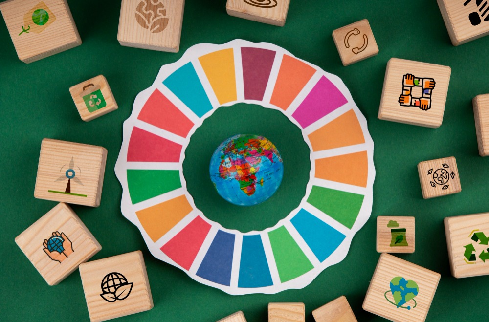 A imagem mostra uma representação dos **Objetivos de Desenvolvimento Sustentável (ODS)** das Nações Unidas. No centro, há um pequeno globo terrestre, simbolizando o planeta, cercado pelo círculo colorido dos 17 ODS. Ao redor, blocos de madeira com ícones relacionados à **sustentabilidade**, como reciclagem, energia limpa, preservação ambiental e cooperação, reforçam a ideia de ações integradas para um futuro mais sustentável. O fundo verde complementa o tema ecológico da composição.